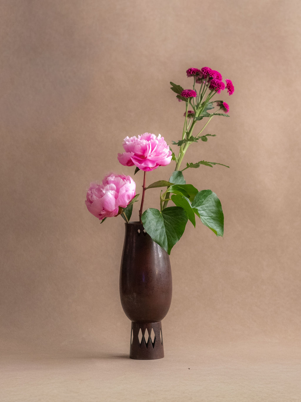 Japanese cast copper vase used with pink flowers, showing its narrow mouth, dark patina, and pierced foot.