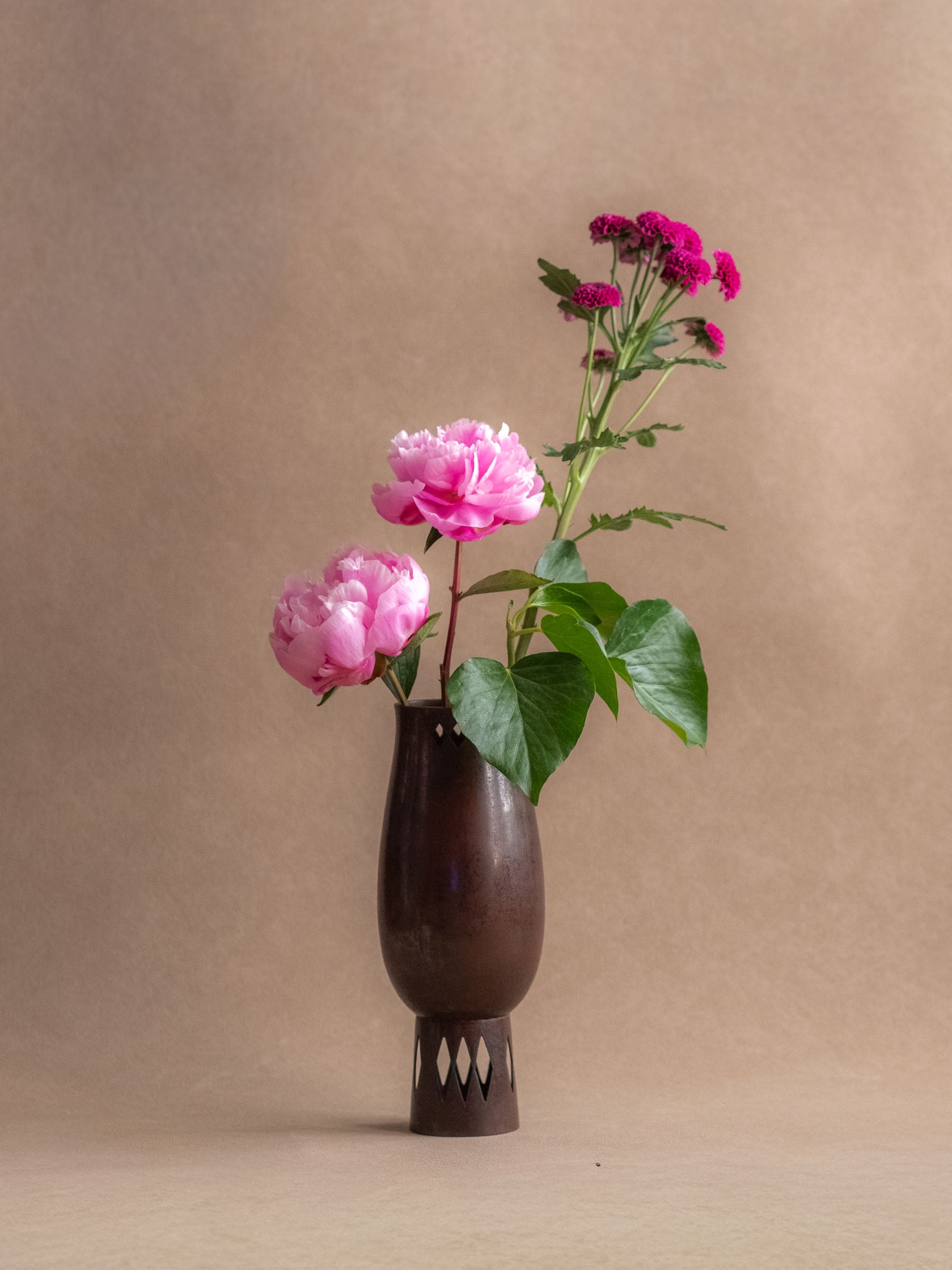 Japanese cast copper vase used with pink flowers, showing its narrow mouth, dark patina, and pierced foot.