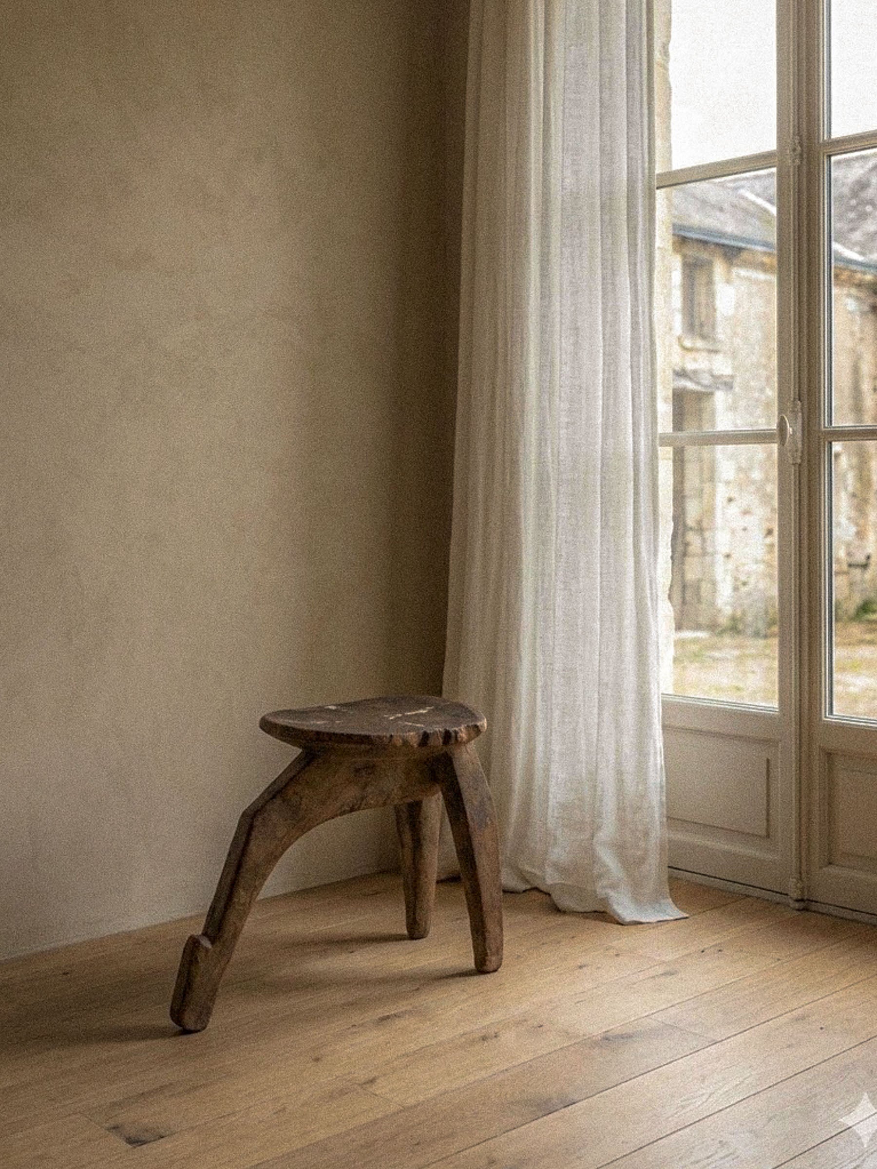 Carved wood Lobi stool with a three-leg construction and aged patina, photographed on a wooden floor beside a tall window with a sheer curtain.