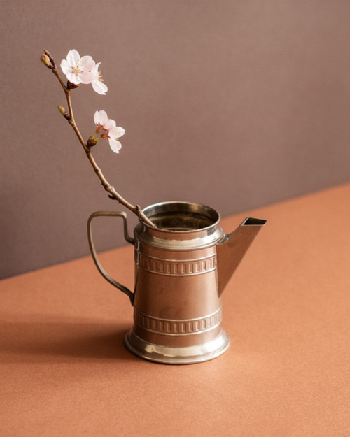 Small silver-plated cream jug on a warm brown background with a cherry blossom branch, shown from the front with tapering body, flared foot, and square-edged spout.