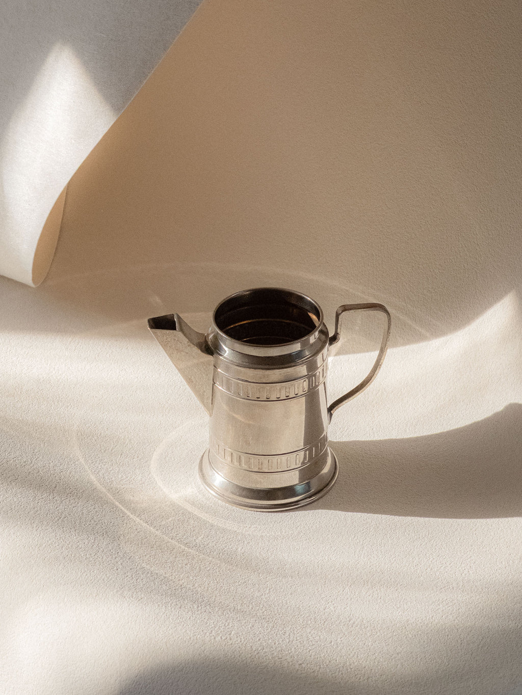 Small silver-plated cream jug photographed front-on in warm natural light, showing the angular spout, strap handle, and banded geometric detail.