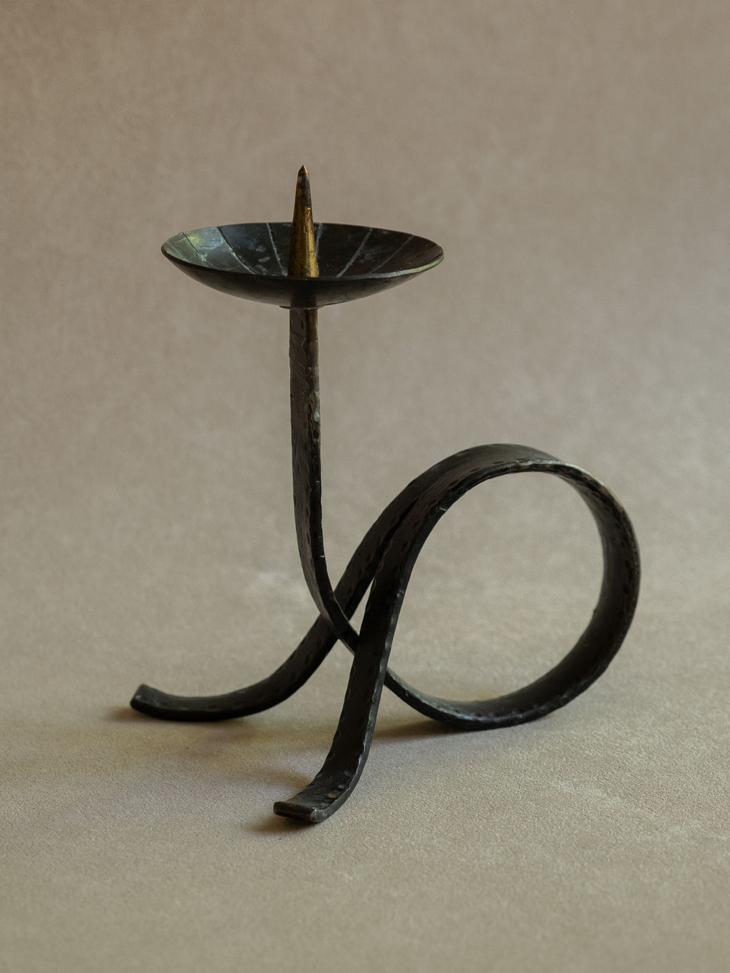 Three-quarter view of the wrought iron candleholder showing the raised pricket, hammered upright stem, circular loop base, and dark patinated surface.