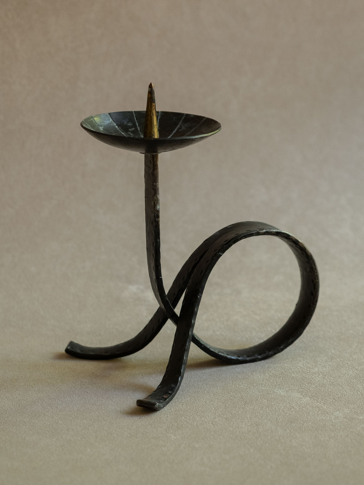 Three-quarter view of the wrought iron candleholder showing the raised pricket, hammered upright stem, circular loop base, and dark patinated surface.