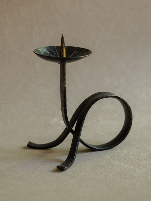 Three-quarter view of the wrought iron candleholder showing the raised pricket, hammered upright stem, circular loop base, and dark patinated surface.