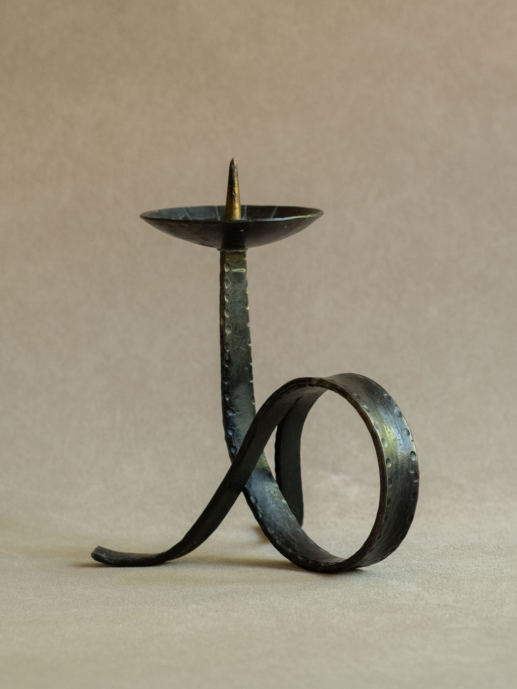 Side view of the wrought iron candleholder with a large looped base, curved foot, blackened metal surface, and brass-toned central spike.