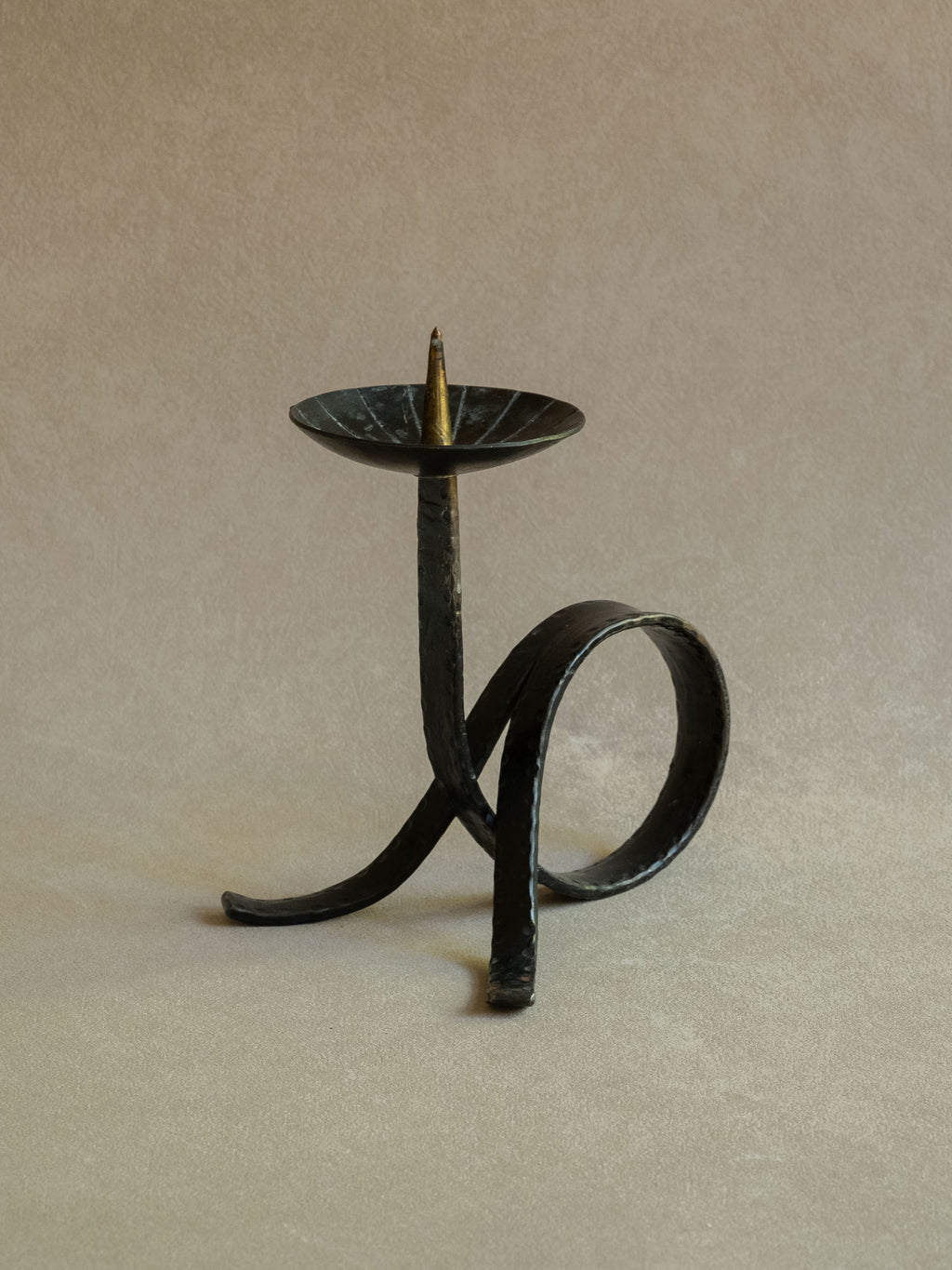 Angled view of the wrought iron candleholder showing the looped strip-iron base, shallow top dish, central pricket, and simple sculptural form.
