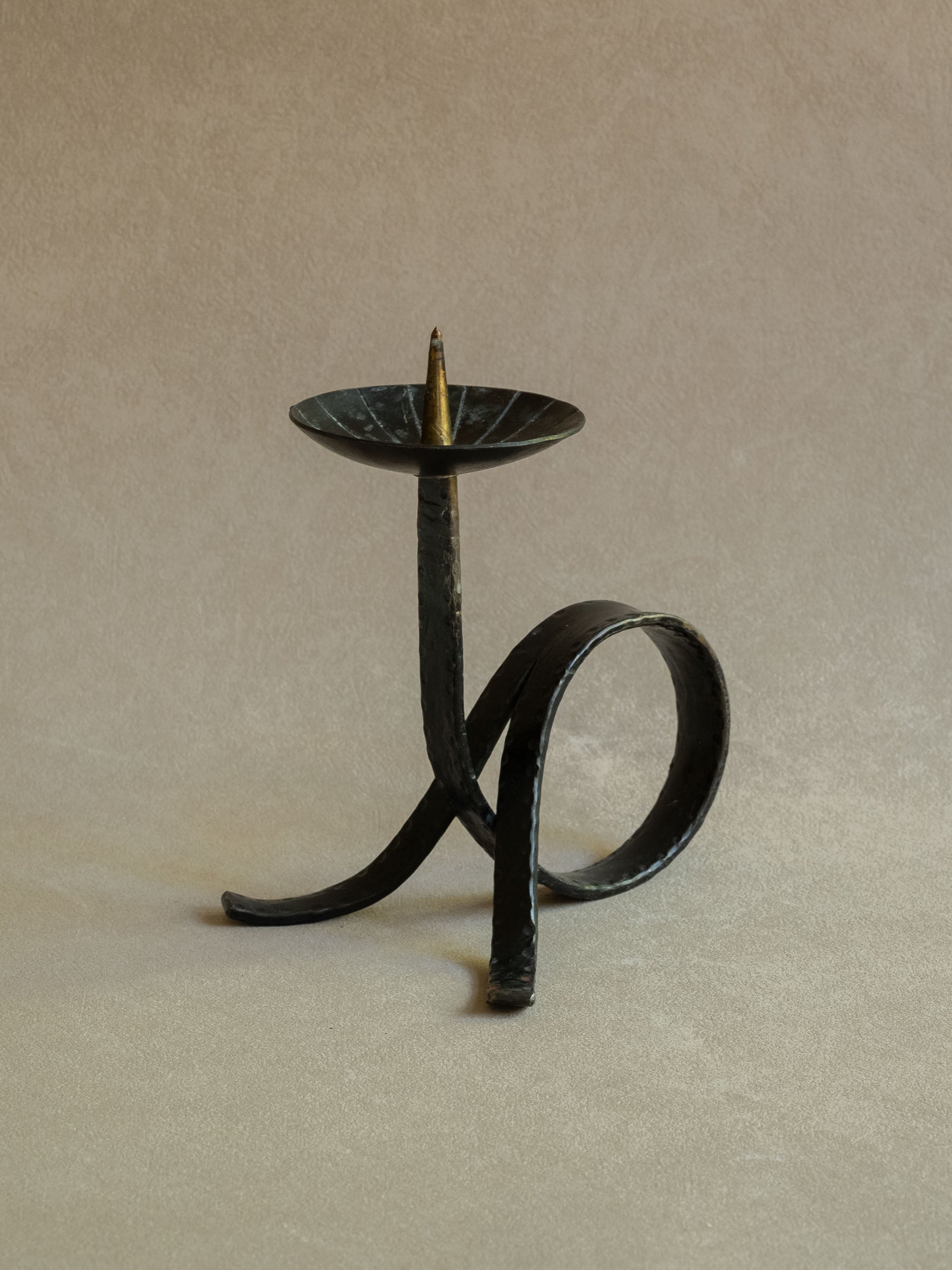 Angled view of the wrought iron candleholder showing the looped strip-iron base, shallow top dish, central pricket, and simple sculptural form.