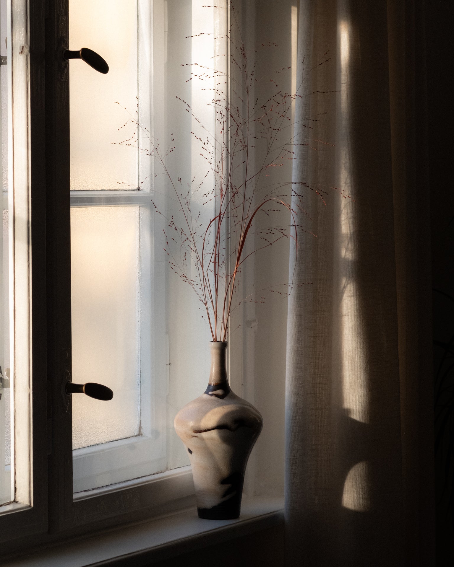 Bulbous ceramic vase with dried grasses on a windowsill in soft window light.