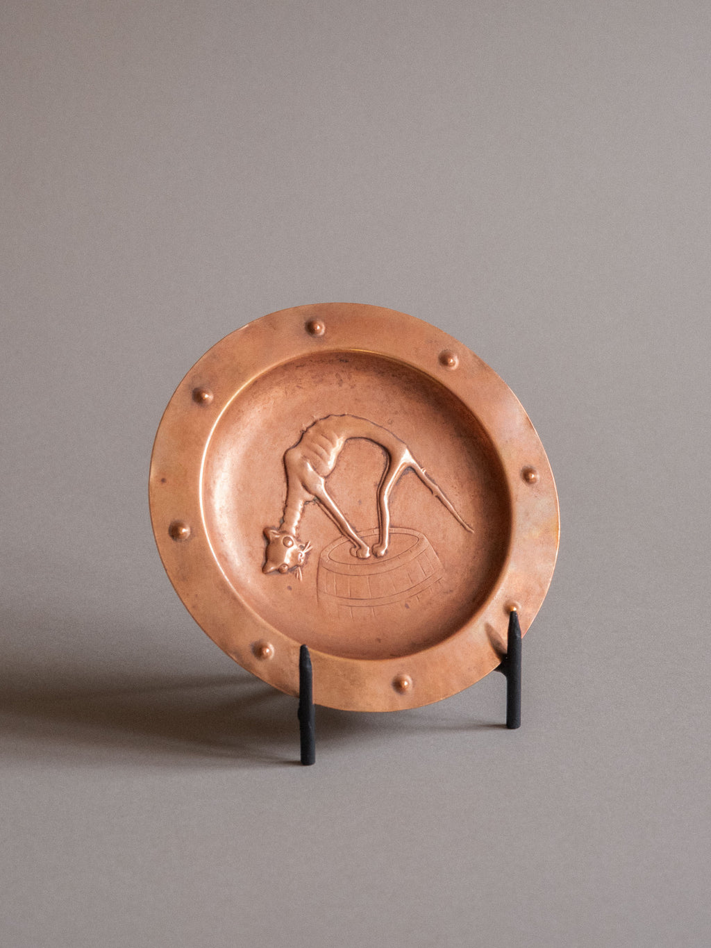 Three-quarter view of a circular copper repoussé plate showing the cat motif, shallow well, and bossed rim.