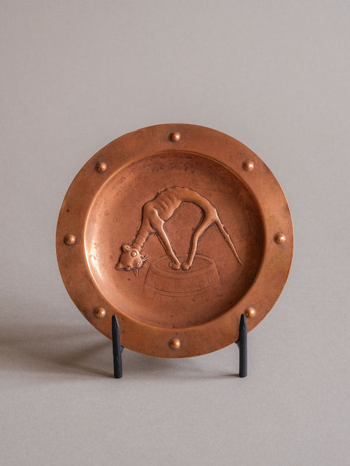 Front view of a small copper repoussé plate with raised cat and barrel motif and domed boss border.