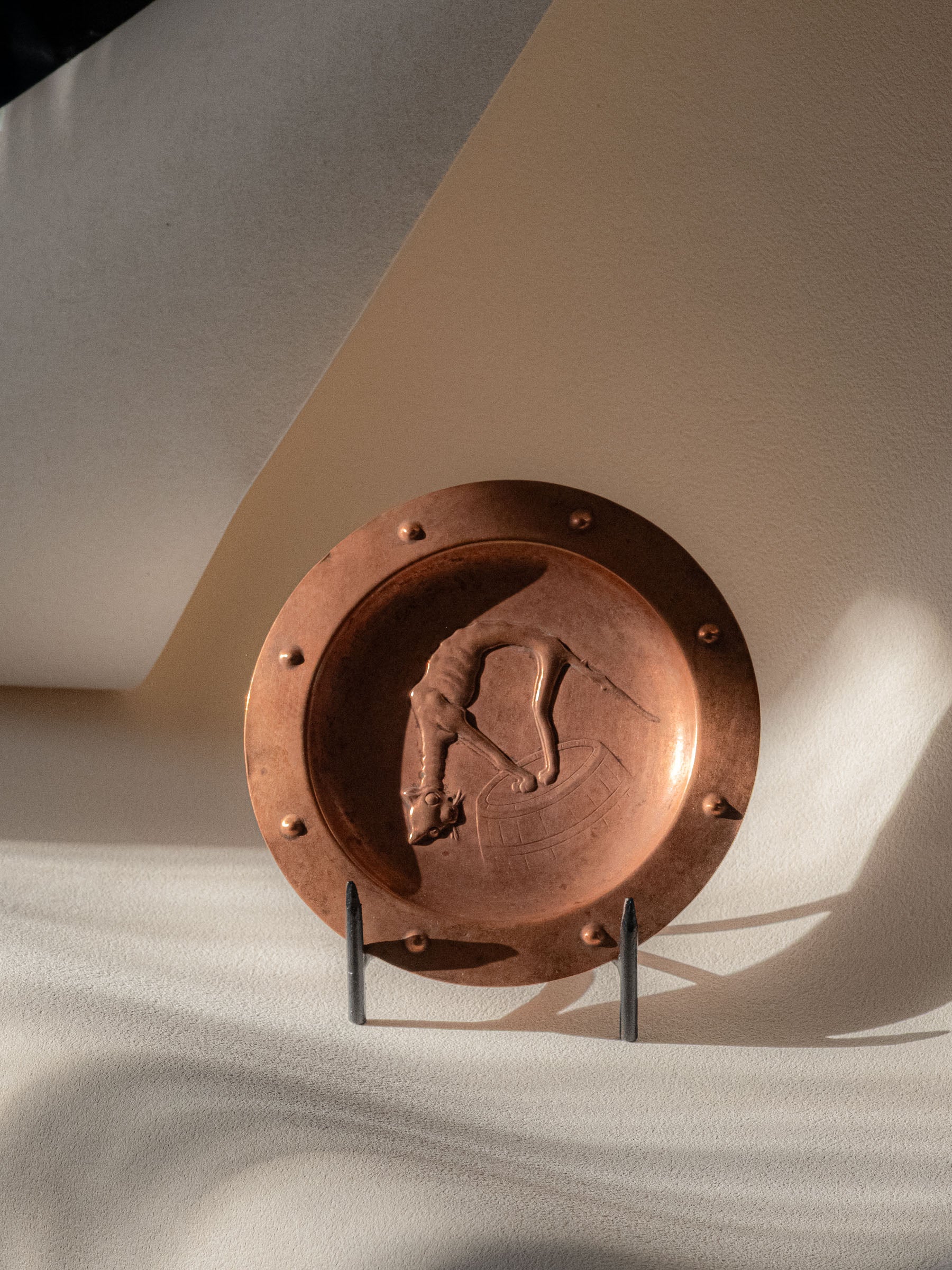 Copper repoussé cat and barrel plate photographed on a stand in angled natural light.
