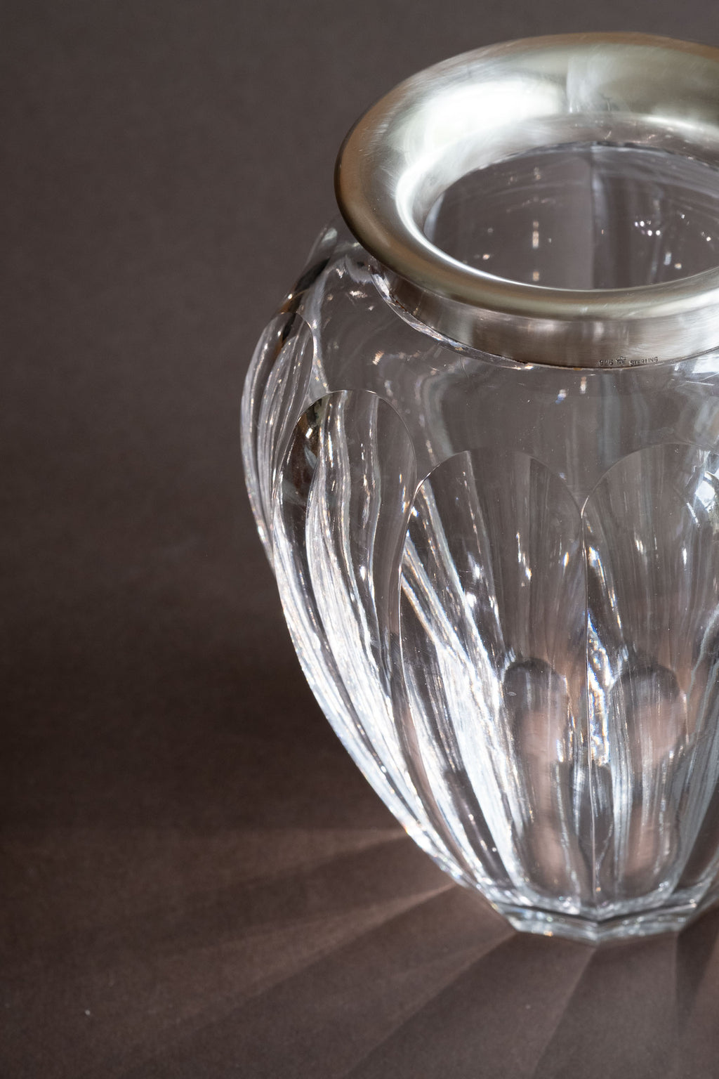 Angled close-up of the crystal vase showing the wide silver rim, cut glass body and light reflected through the surface.
