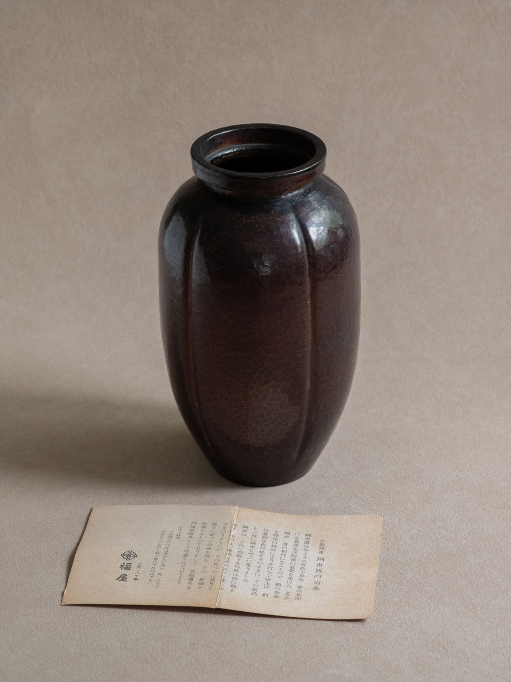 Full view of a Japanese Dochu hand-hammered copper vase with rounded ribbed body, raised rim, and Fukuya paper insert below.