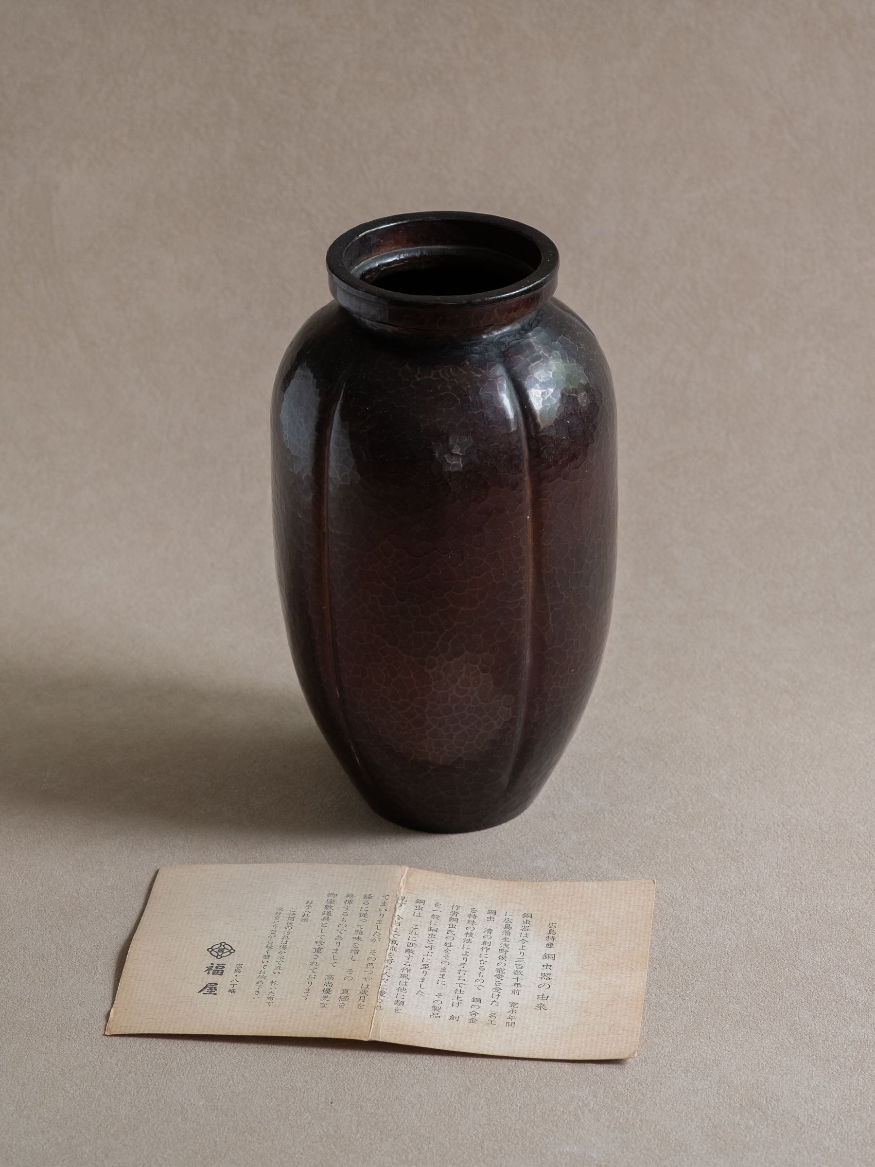 Full view of a Japanese Dochu hand-hammered copper vase with rounded ribbed body, raised rim, and Fukuya paper insert below.