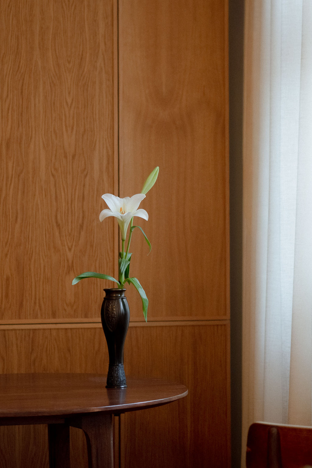Japanese bronze flower vase by Kanamori Shōei shown with a while Lily flower, featuring a tall tapered form and relief panel detail.