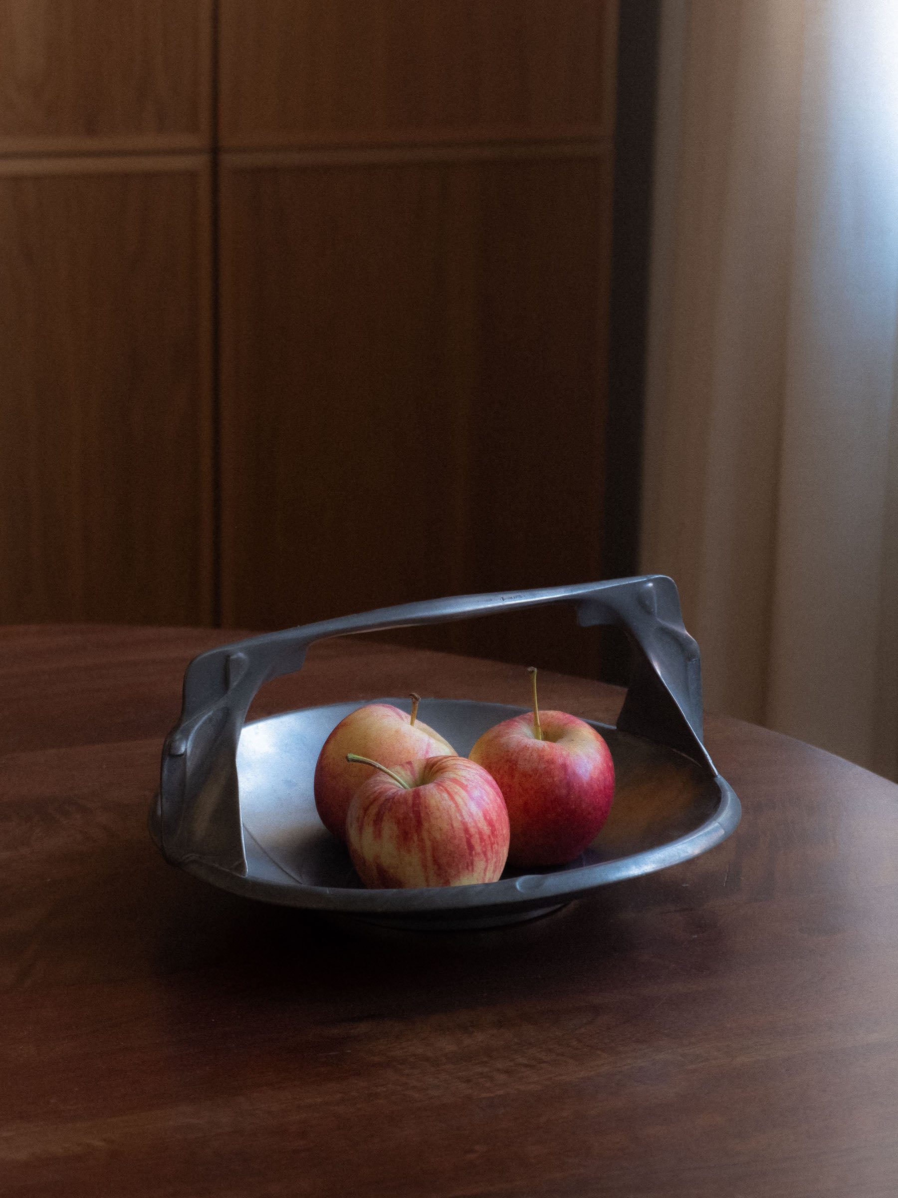 Dark metal handled fruit bowl on a round wooden table, holding three red apples, with warm wood cabinetry and soft window light in the background.