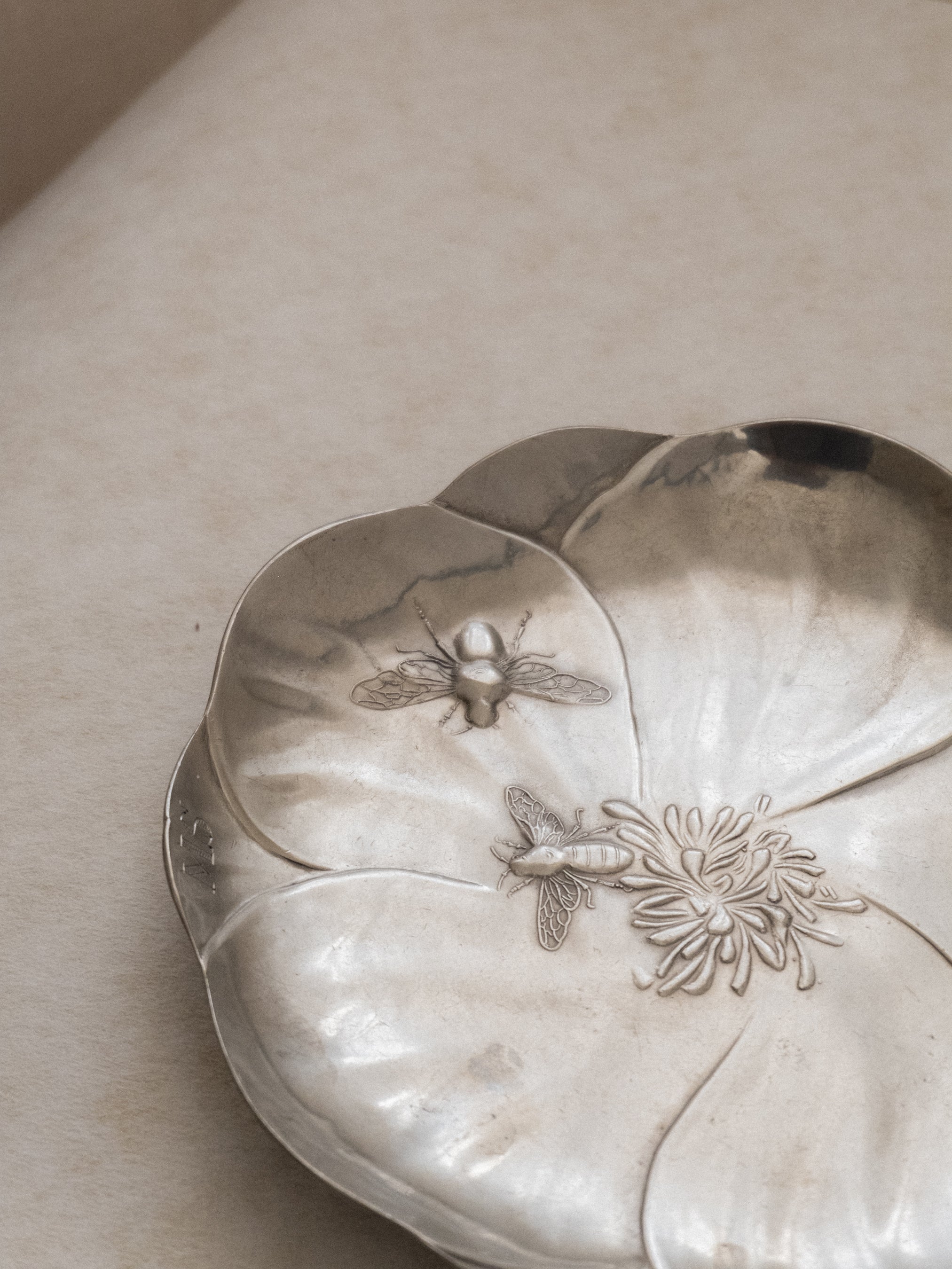 Angled top view of the pewter bee dish lying flat, showing the flower-shaped basin and relief insect decoration.