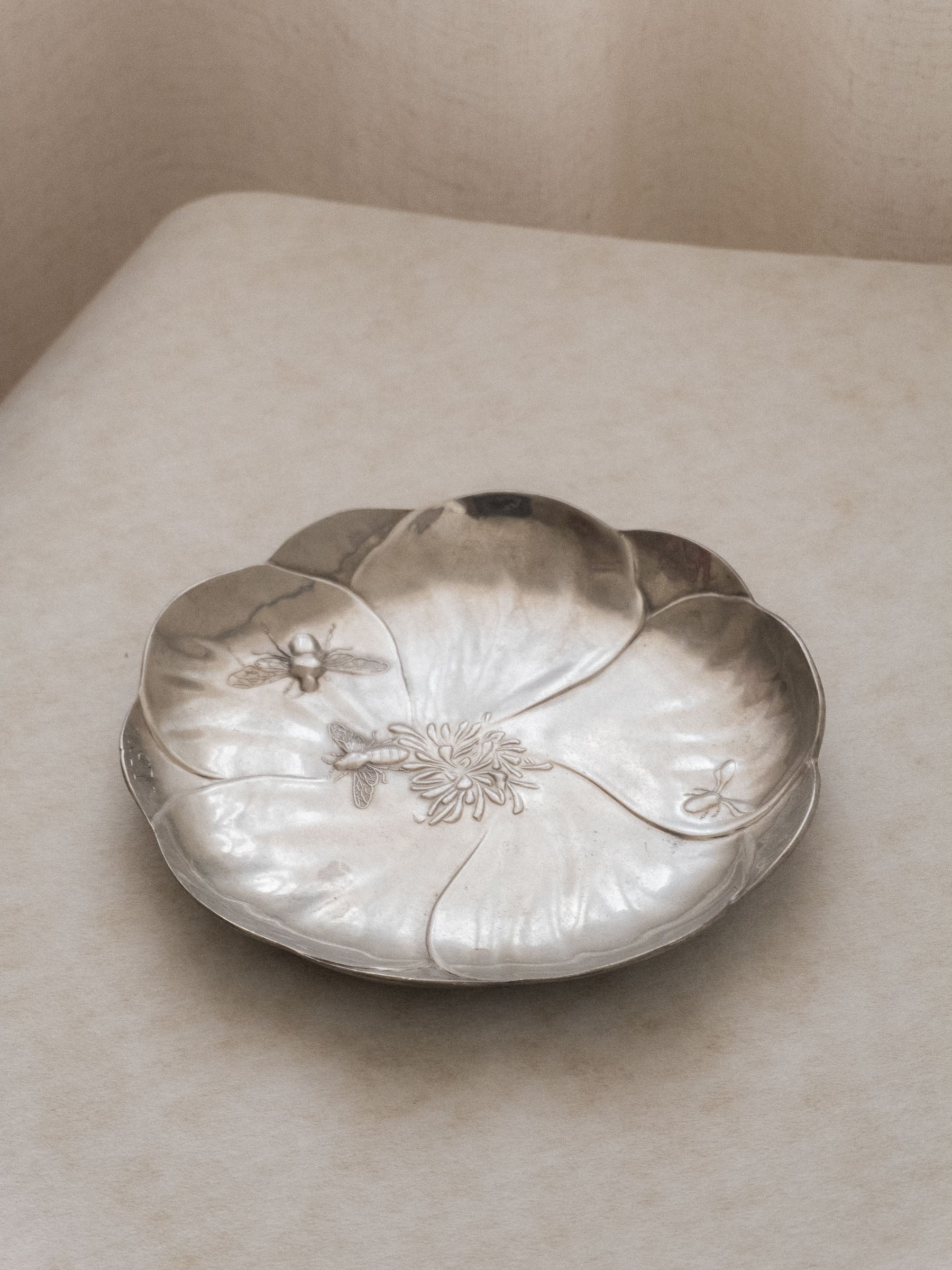 Angled top view of the pewter bee dish lying flat, showing the flower-shaped basin and relief insect decoration.