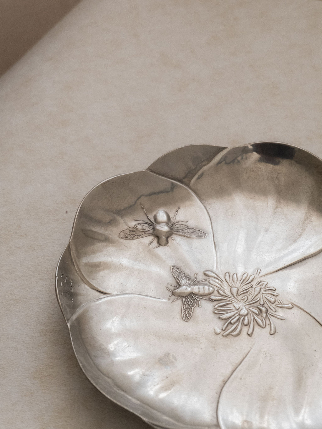 Angled top view of the pewter bee dish lying flat, showing the flower-shaped basin and relief insect decoration.