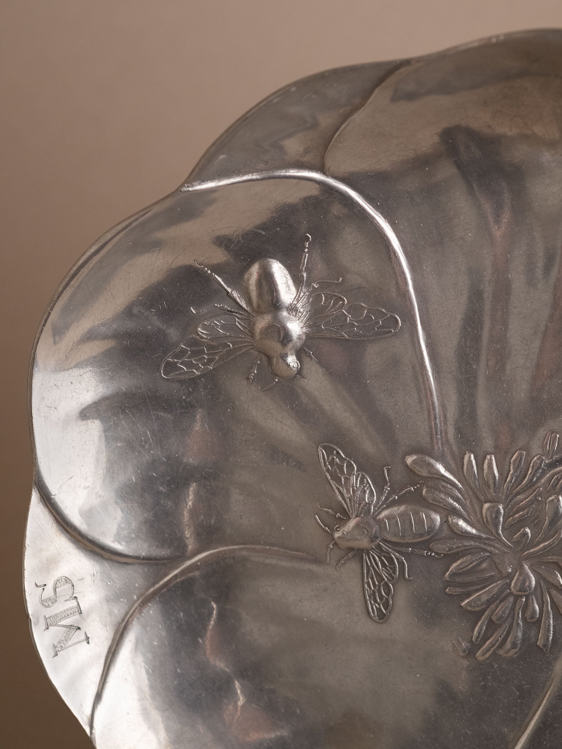 Close-up of two raised bees and the engraved monogram on the rim of the pewter flower dish.