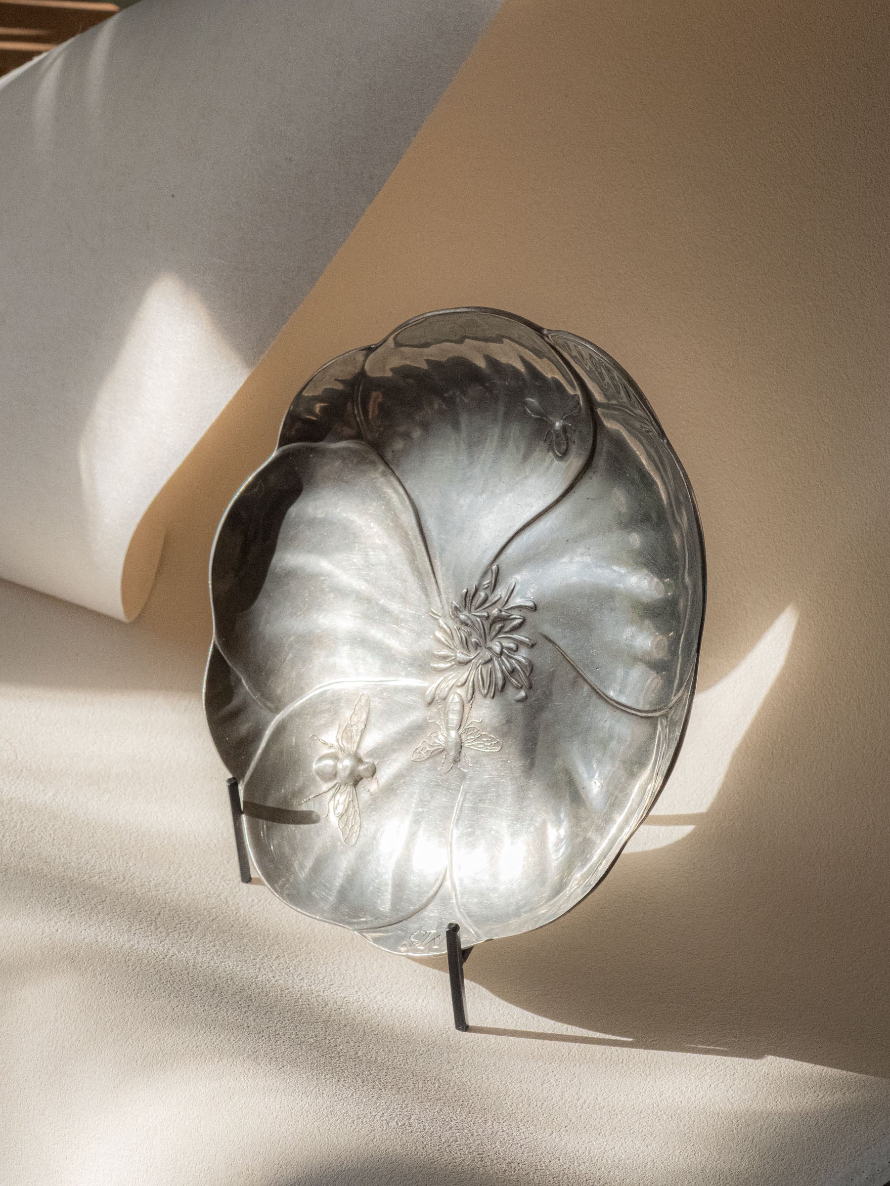 Kayserzinn pewter bee dish displayed upright on a black stand, showing the flower-shaped form and relief bees in angled natural light.