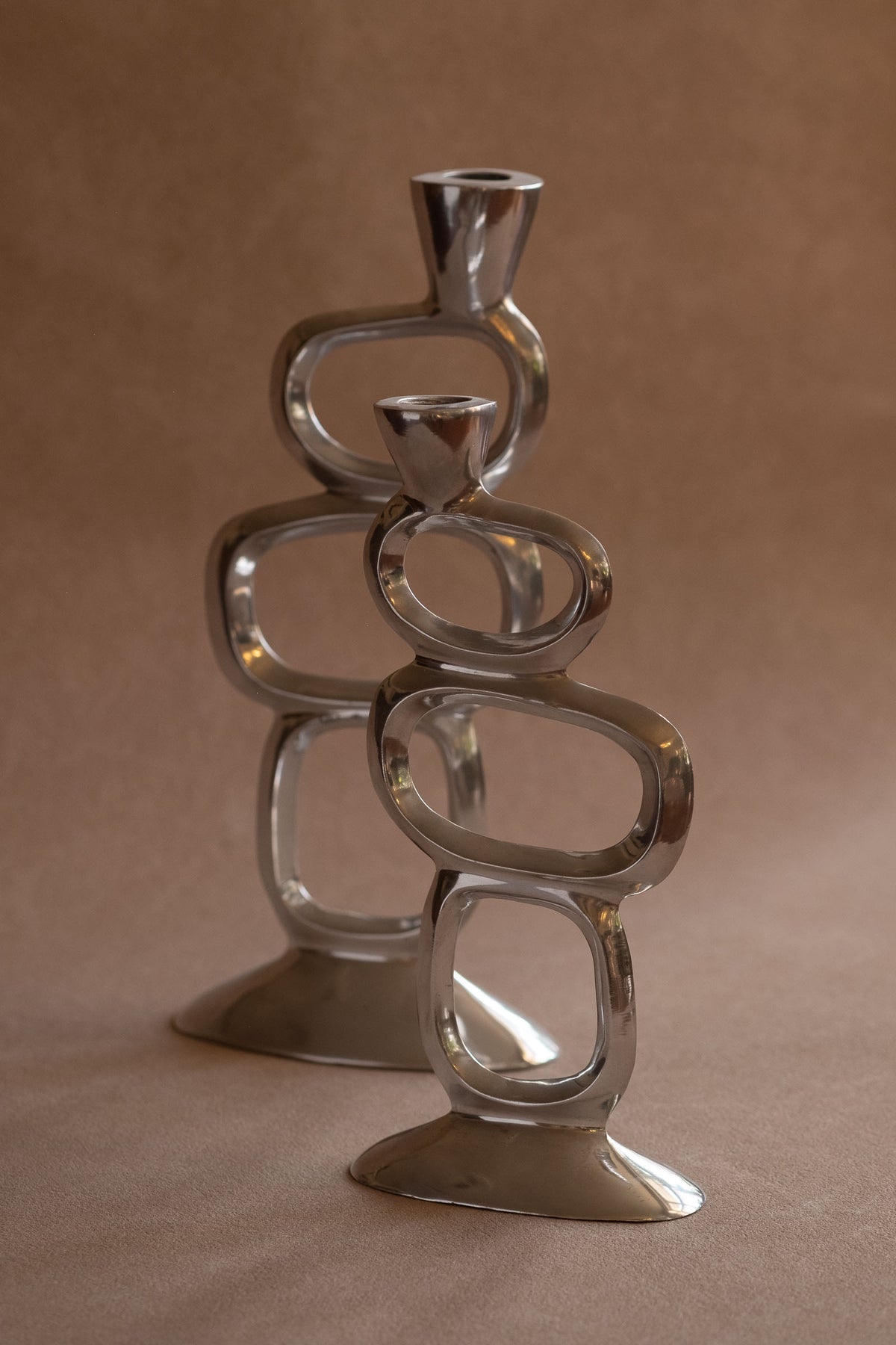 Pair of Matthew Hilton Circle candle holders in polished aluminium, photographed at an angle with the smaller holder in front.

