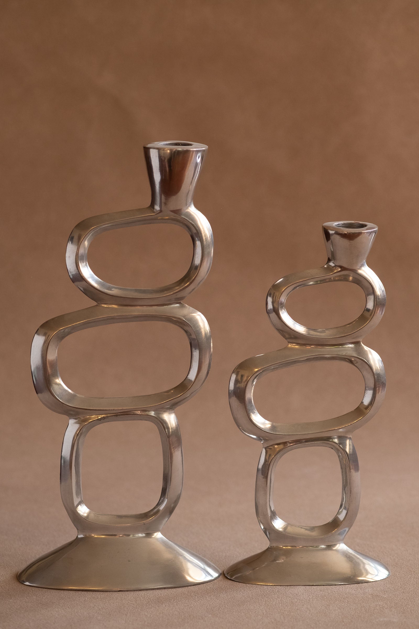 Pair of polished aluminium Matthew Hilton Circle candle holders shown front-facing, with open geometric ring forms and wide bases.
