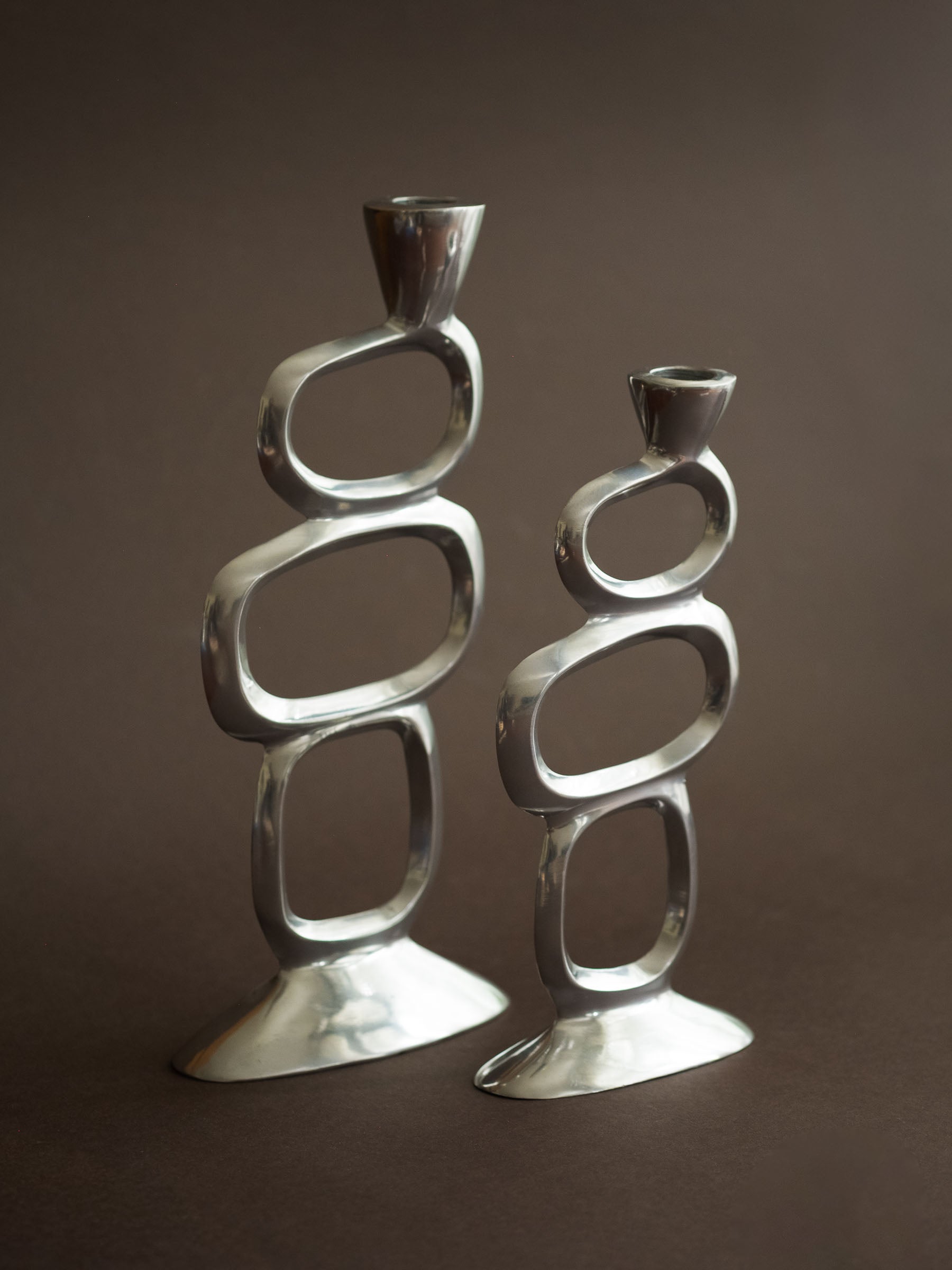 Pair of Matthew Hilton Circle candle holders in polished aluminium, shown from a side angle against a dark brown background.
