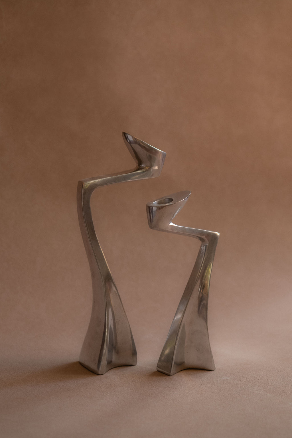 Pair of polished aluminium Swan candle holders shown front-on, with curved upright forms against a warm brown backdrop.
