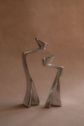 Pair of polished aluminium Swan candle holders shown front-on, with curved upright forms against a warm brown backdrop.
