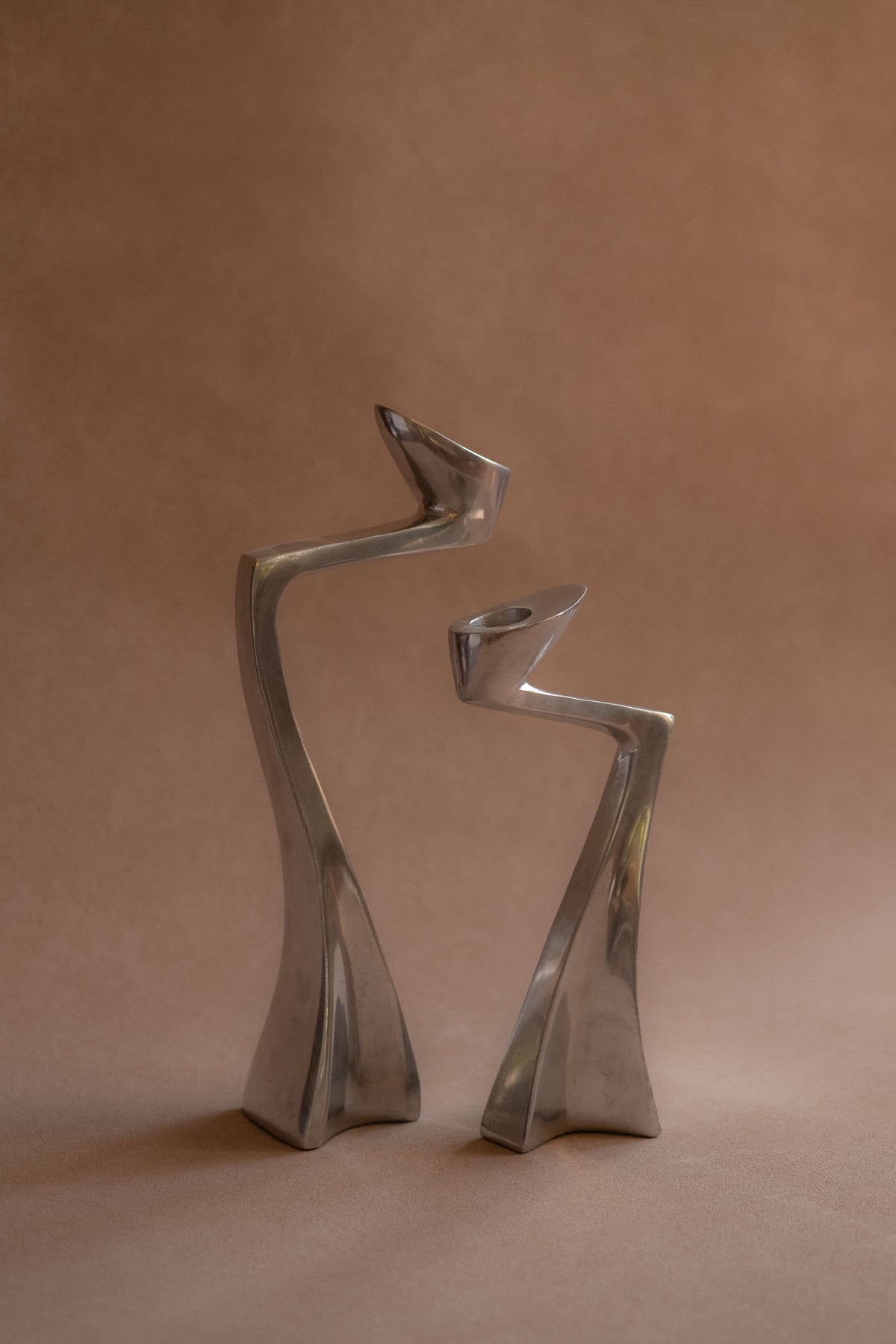 Pair of polished aluminium Swan candle holders shown front-on, with curved upright forms against a warm brown backdrop.
