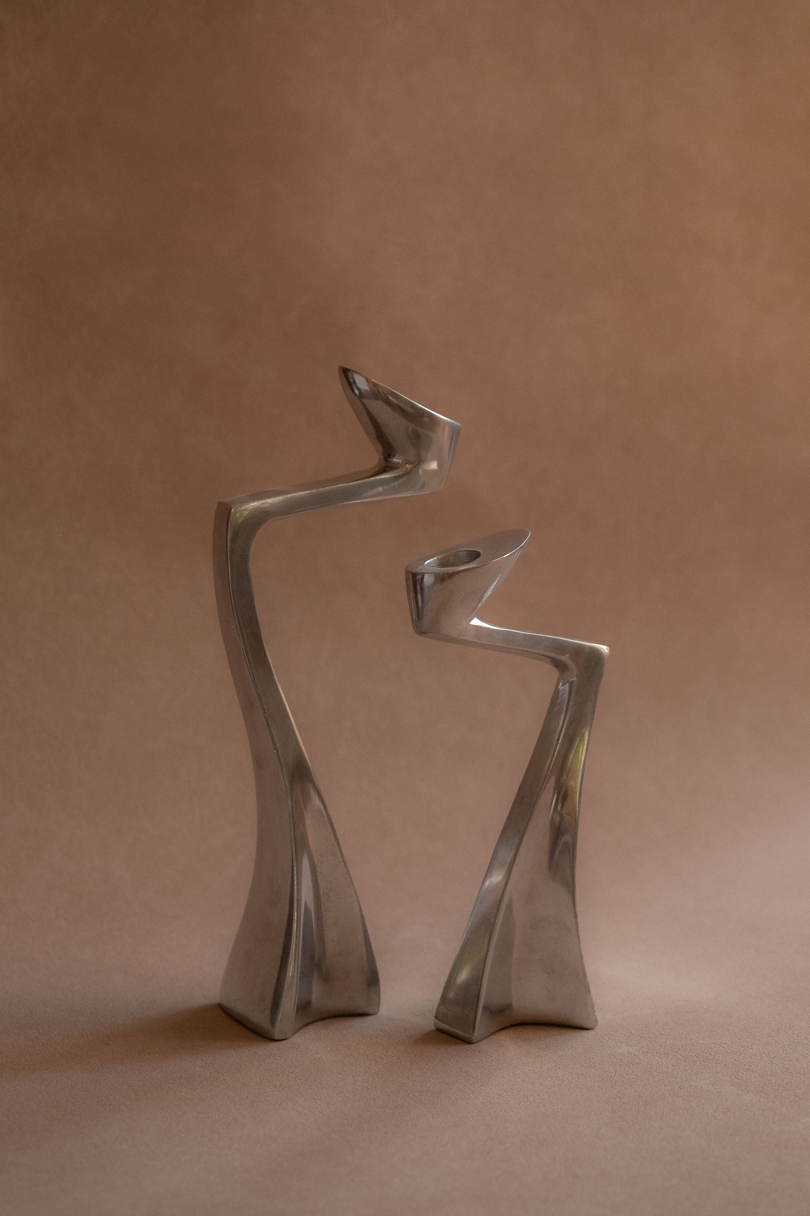 Pair of polished aluminium Swan candle holders shown front-on, with curved upright forms against a warm brown backdrop.

