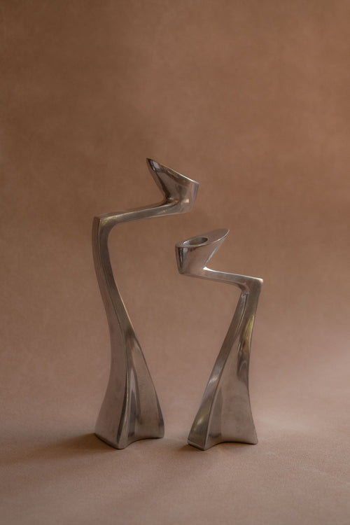 Pair of polished aluminium Swan candle holders shown front-on, with curved upright forms against a warm brown backdrop.

