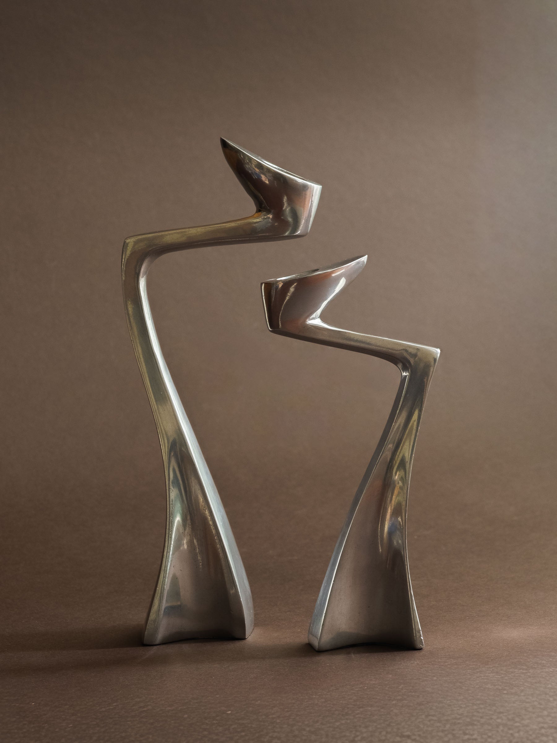 Pair of polished aluminium Swan candle holders shown front-on, with curved profiles and reflective silver-toned surfaces.
