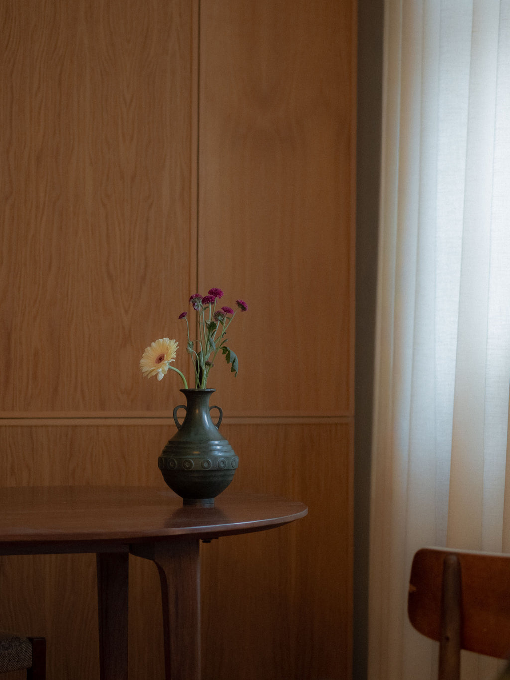 Murata Yoshio Janome bronze vase, Japan, Shōwa period, styled with fresh flowers on a wooden table beside a sheer curtain in a warm interior setting.