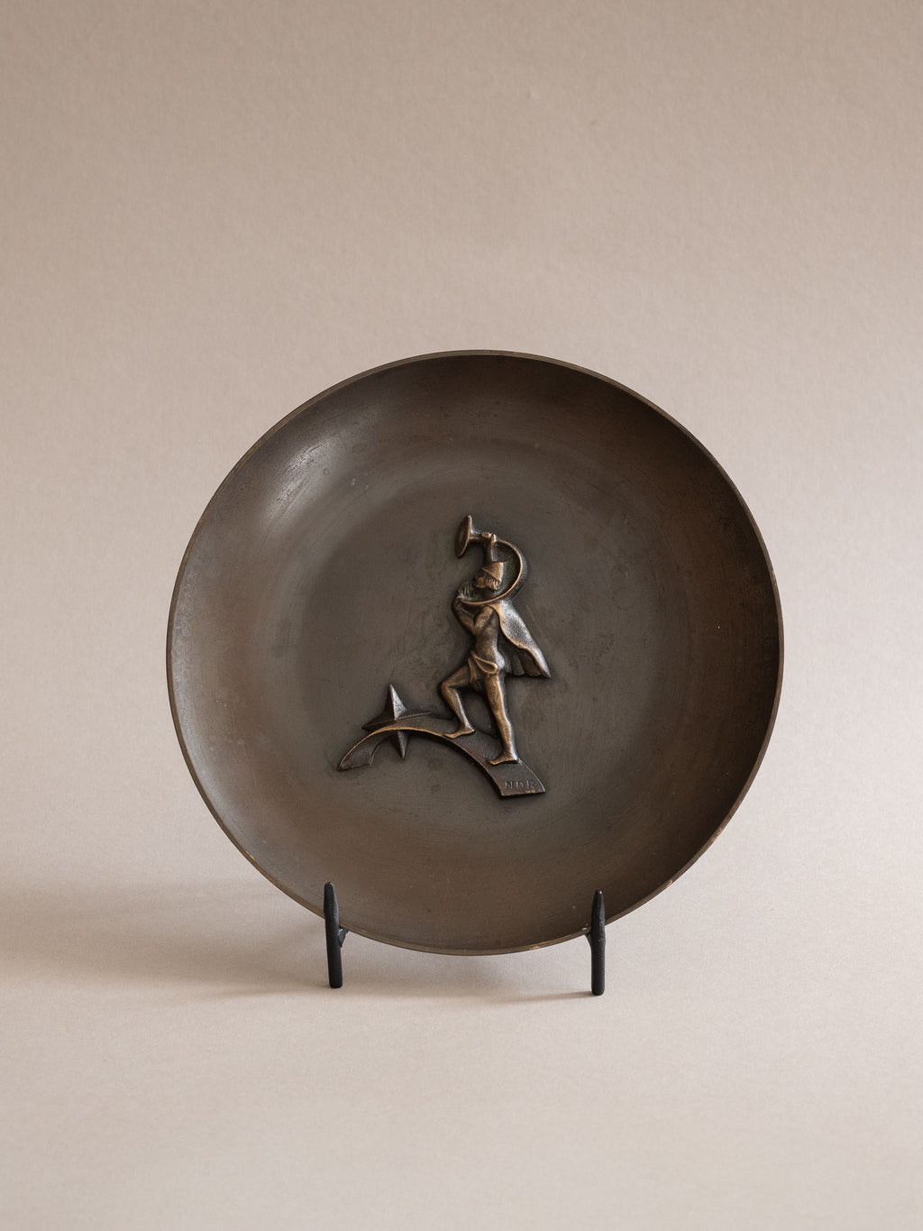 Front view of a Nordisk Malm figural bronze dish showing the shallow circular form and central horn-blower relief, displayed on a black stand.

