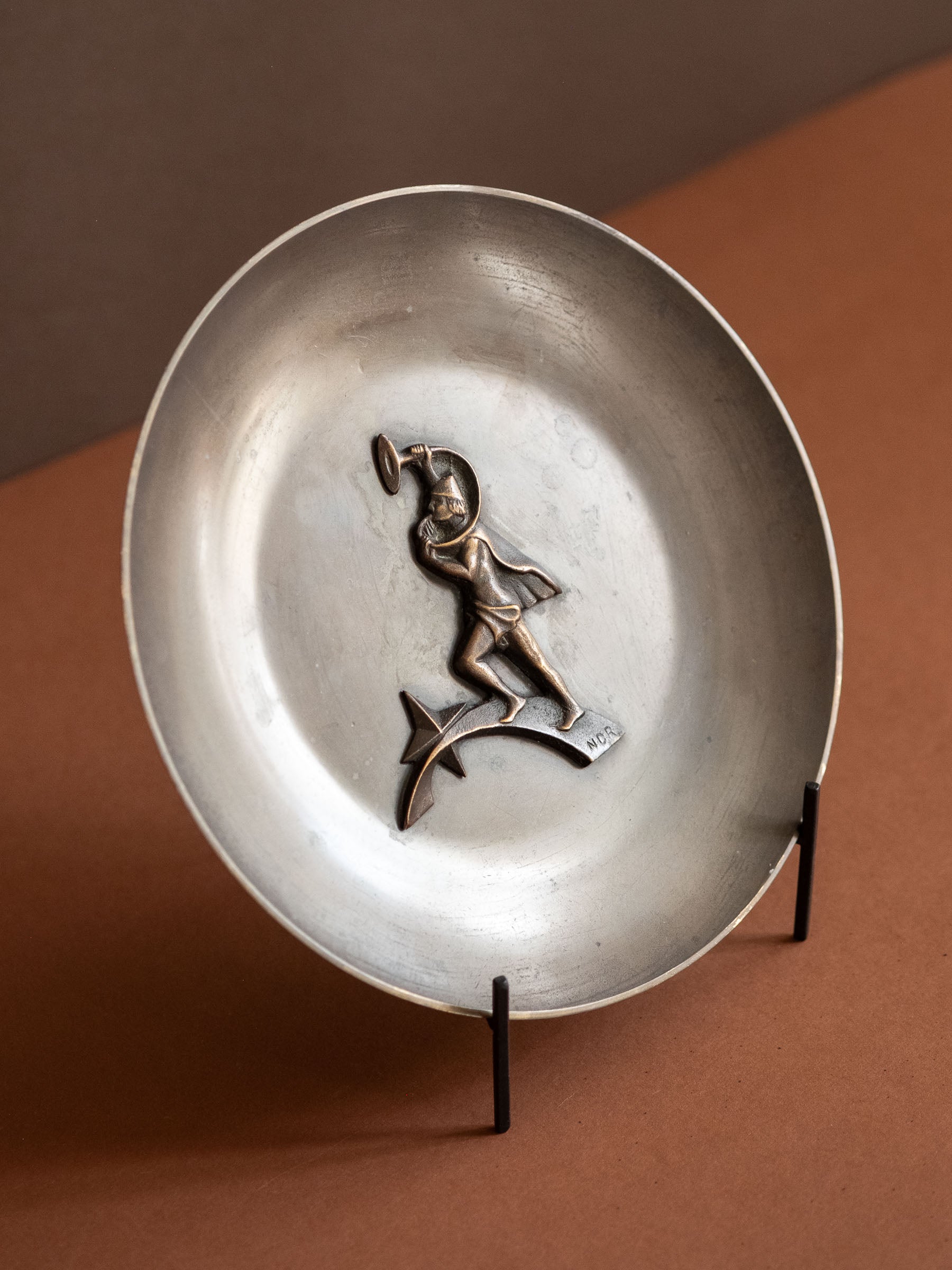 Angled view of a Nordisk Malm figural bronze dish with dark patina and raised horn-blower relief, displayed on a black stand against a warm brown background.
