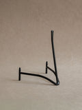 Side view of a black iron plate stand with curved base, front supports, and tall upright back against a warm beige background