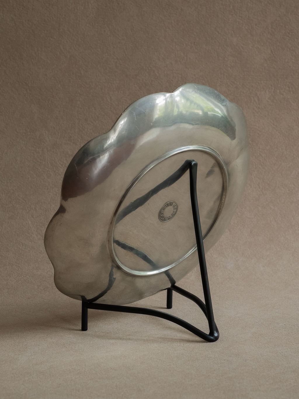 Black iron plate stand holding a scalloped silver-toned tray at a slight angle against a warm beige background