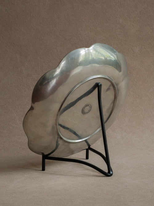 Black iron plate stand holding a scalloped silver-toned tray at a slight angle against a warm beige background