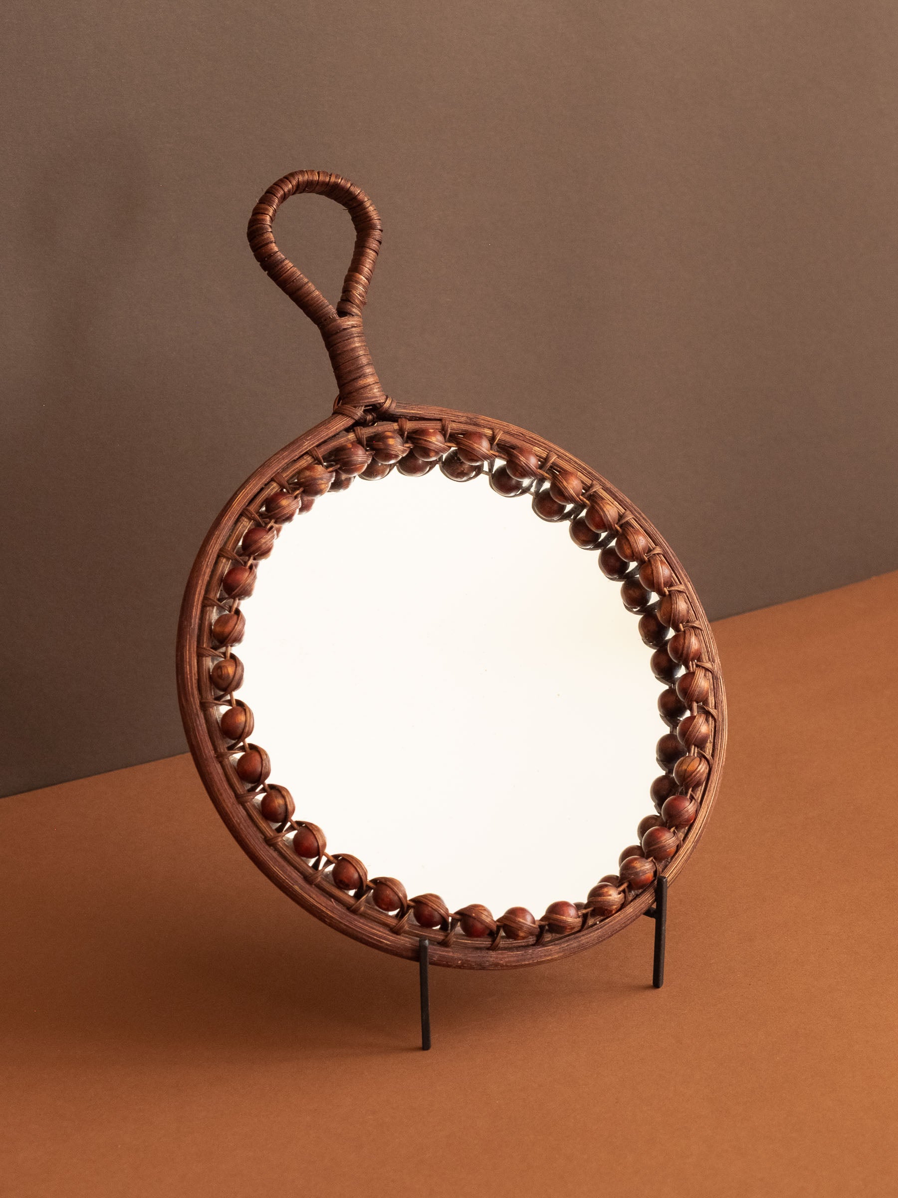 Round beaded rattan wall mirror with loop handle shown at an angle on a stand against a brown background.
