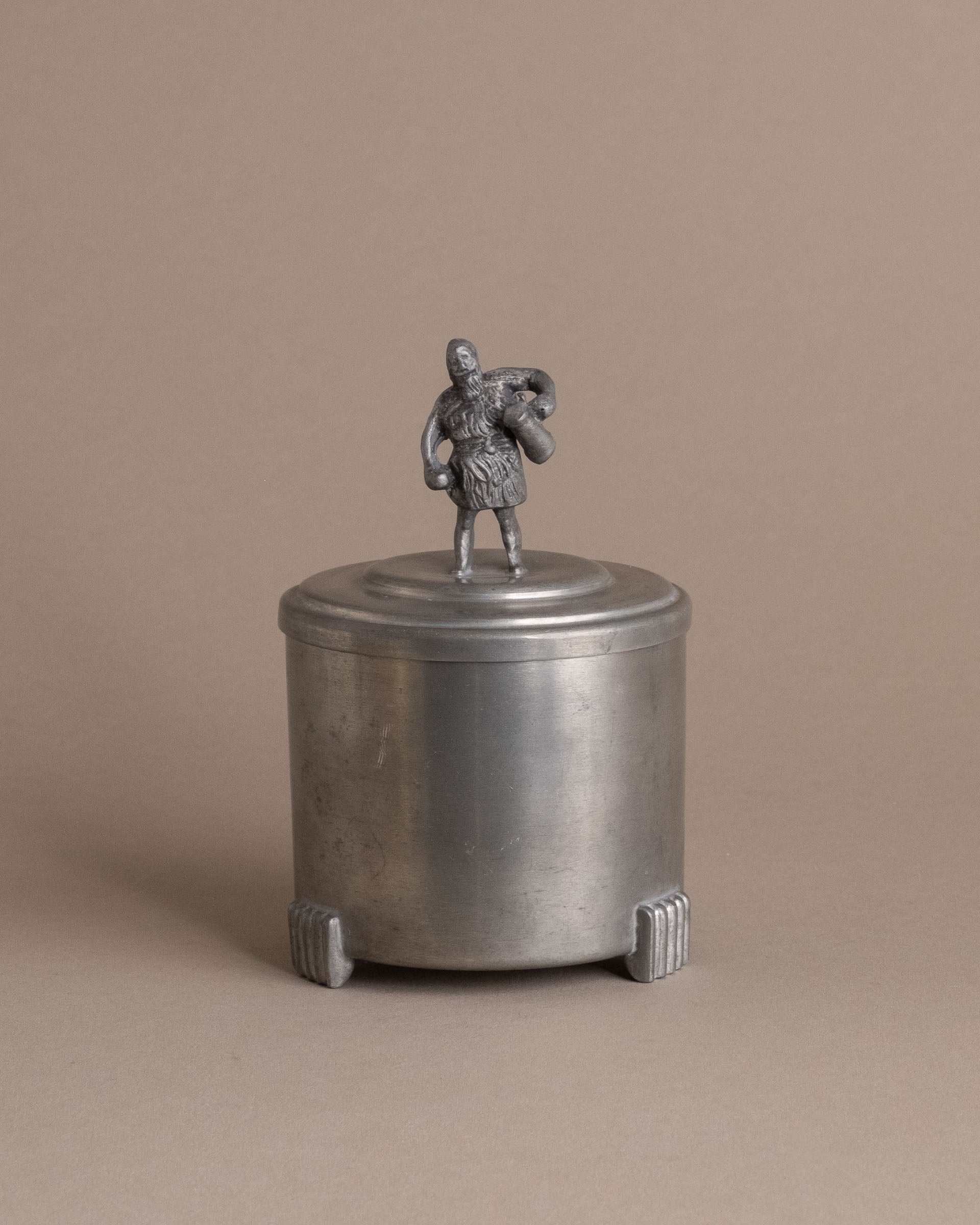 Front view of a small Savo Tinn pewter lidded box with a male figure finial and three ribbed feet.