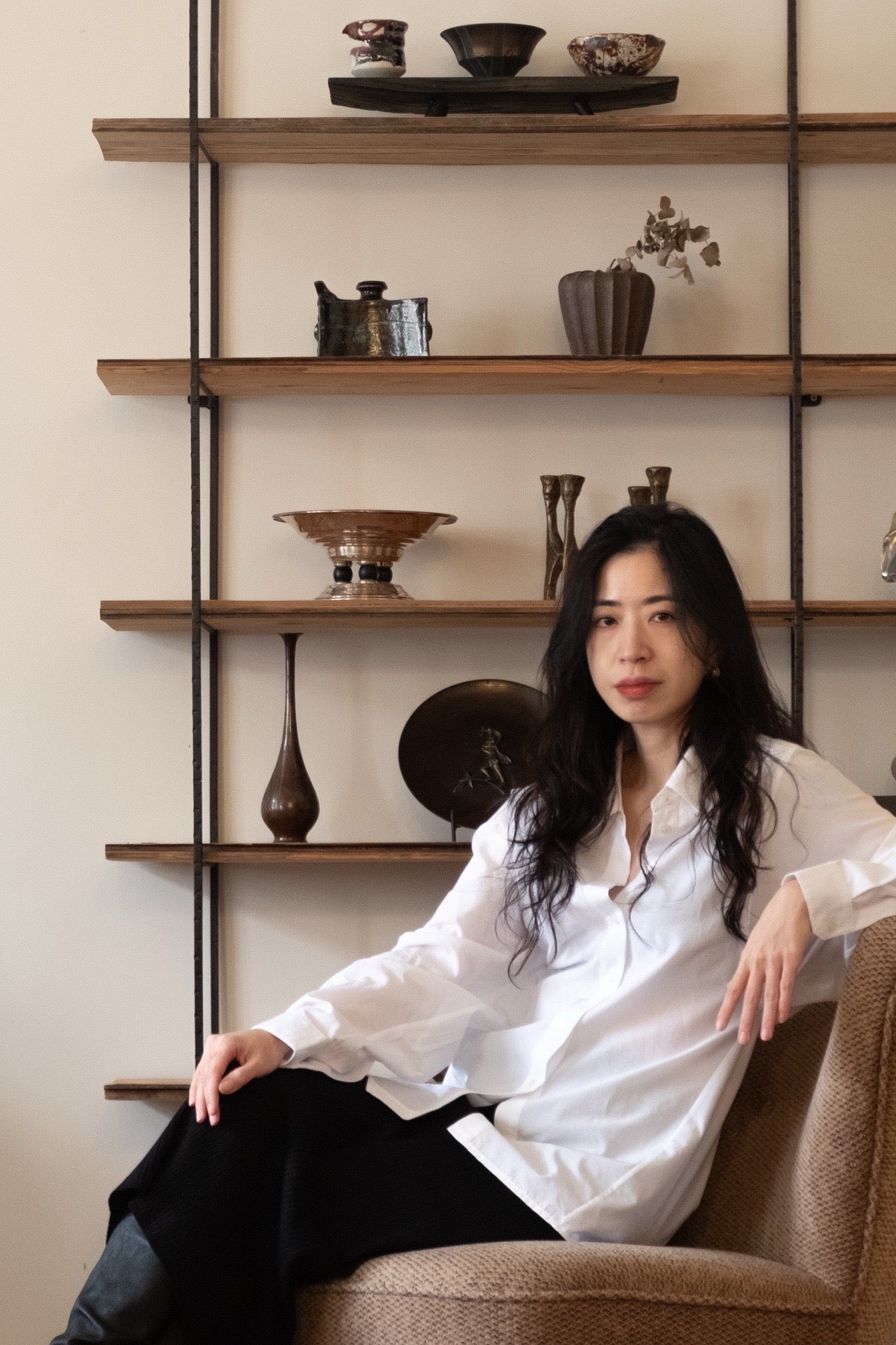 Founder of The Linie seated in a lounge chair in front of wooden shelving displaying curated vintage and antique objects.