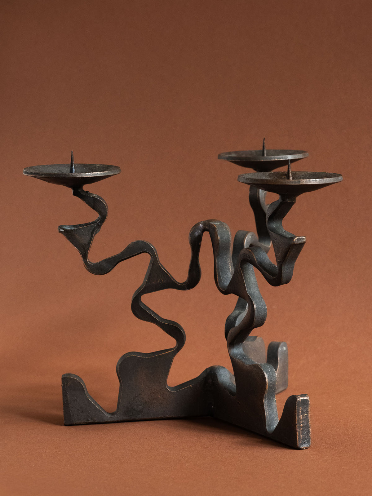 Three-quarter view of a three-arm Brutalist forged iron candleholder with blackened surface, warm copper-toned edges and three pricket dishes.