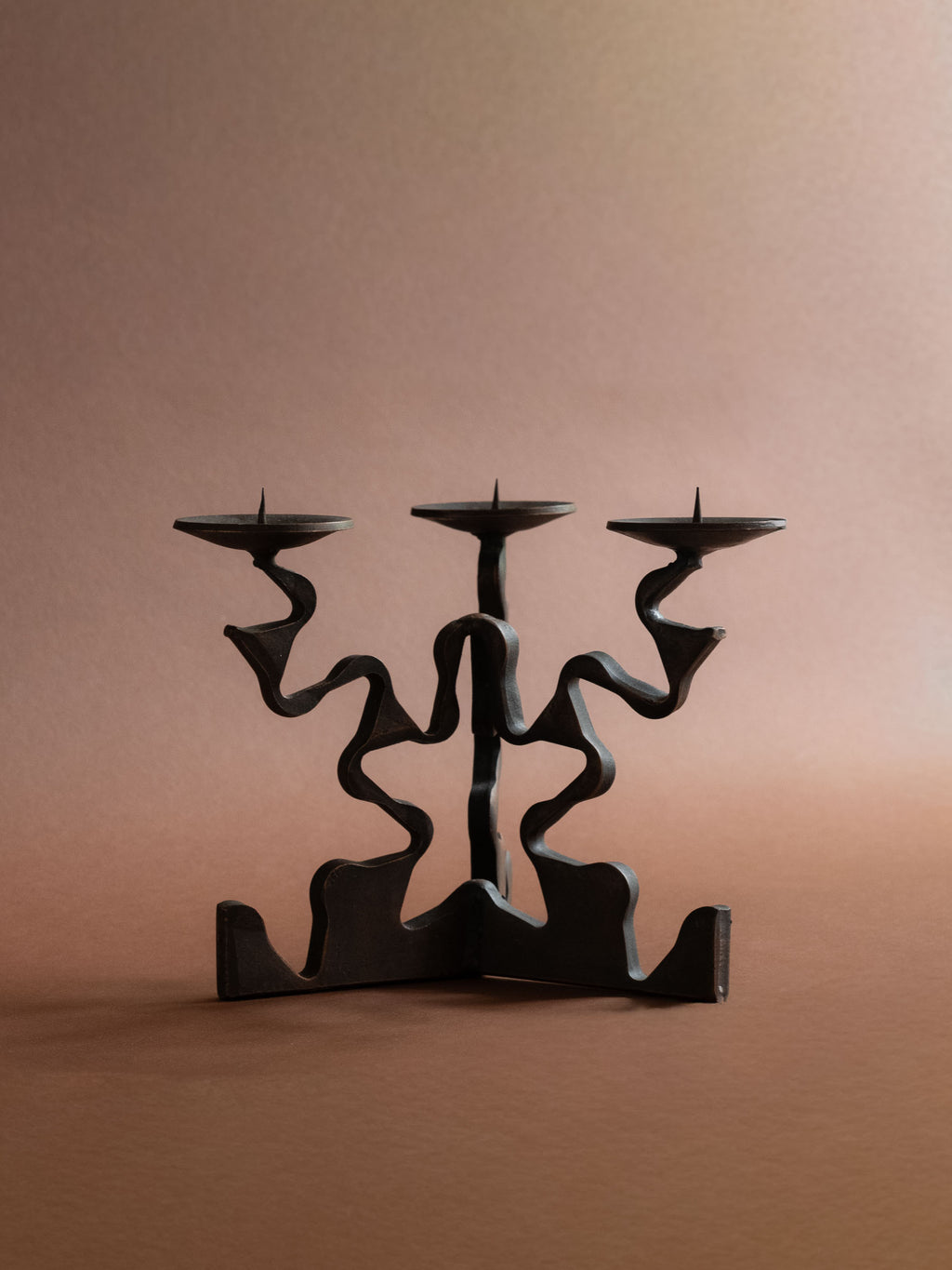 Front view of the forged iron three-arm candleholder showing its symmetrical wave-cut base, looping arms and three pricket candle dishes.