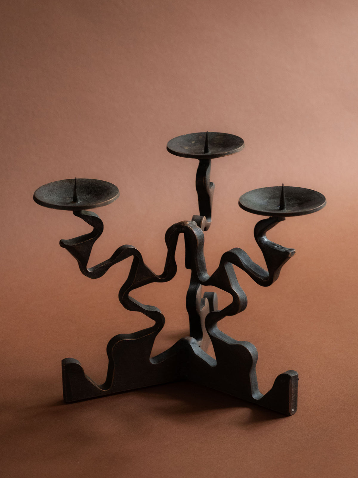 Elevated three-quarter view of the Brutalist candleholder showing three circular pricket dishes and curling ribbon-like forged arms.