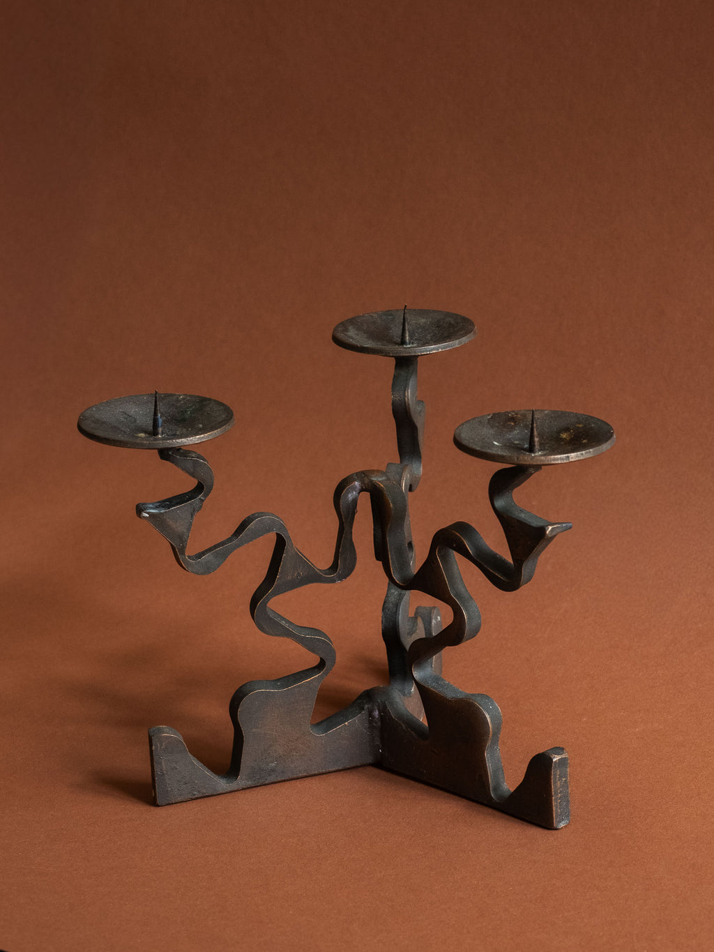 Full view of the forged iron candleholder showing the wave-cut base, strong negative space and warm copper-toned wear along the edges.