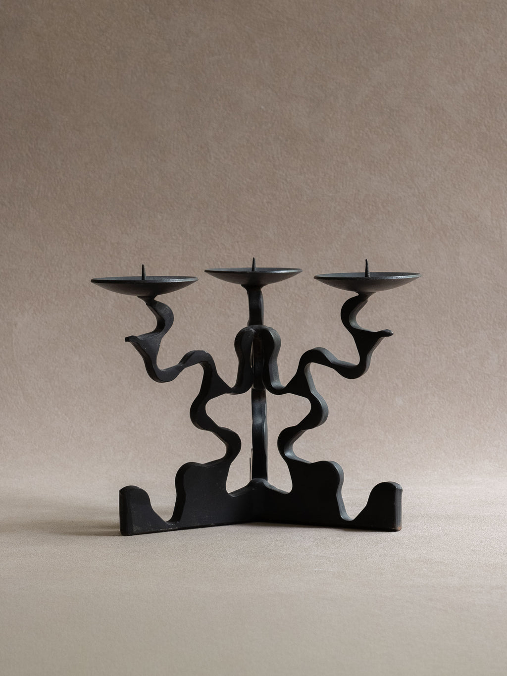 Front view of the three-arm forged iron candleholder, showing three shallow candle dishes, central pricket spikes, wavy arms, and wide base.