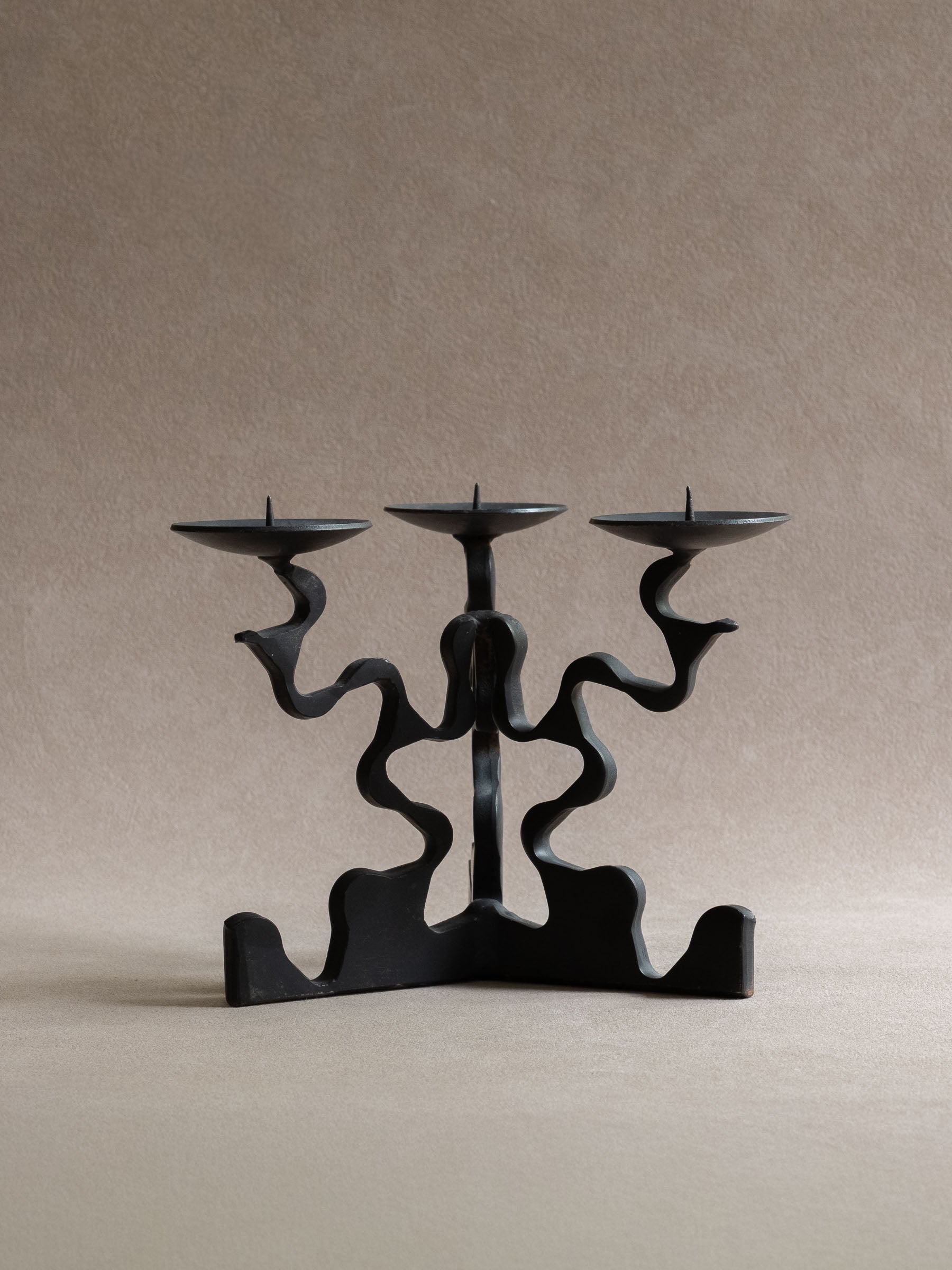Front view of the three-arm forged iron candleholder, showing three shallow candle dishes, central pricket spikes, wavy arms, and wide base.