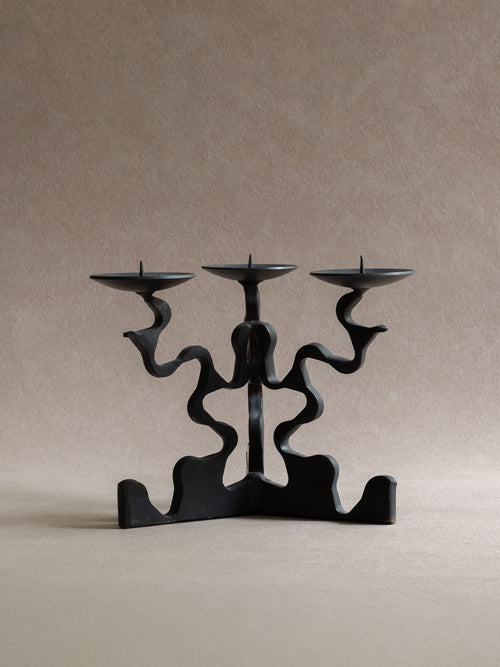 Front view of the three-arm forged iron candleholder, showing three shallow candle dishes, central pricket spikes, wavy arms, and wide base.